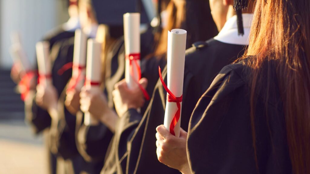 Graduates in black caps and gowns holding diplomas tied with red ribbons, standing in a line, celebrating their graduation ceremony