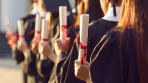 Graduates in black caps and gowns holding diplomas tied with red ribbons, standing in a line, celebrating their graduation ceremony