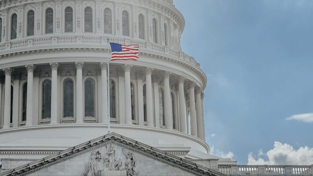 The American flag waves in front of the U.S. Capitol building, featuring its distinctive dome and classical columns against a partly cloudy sky