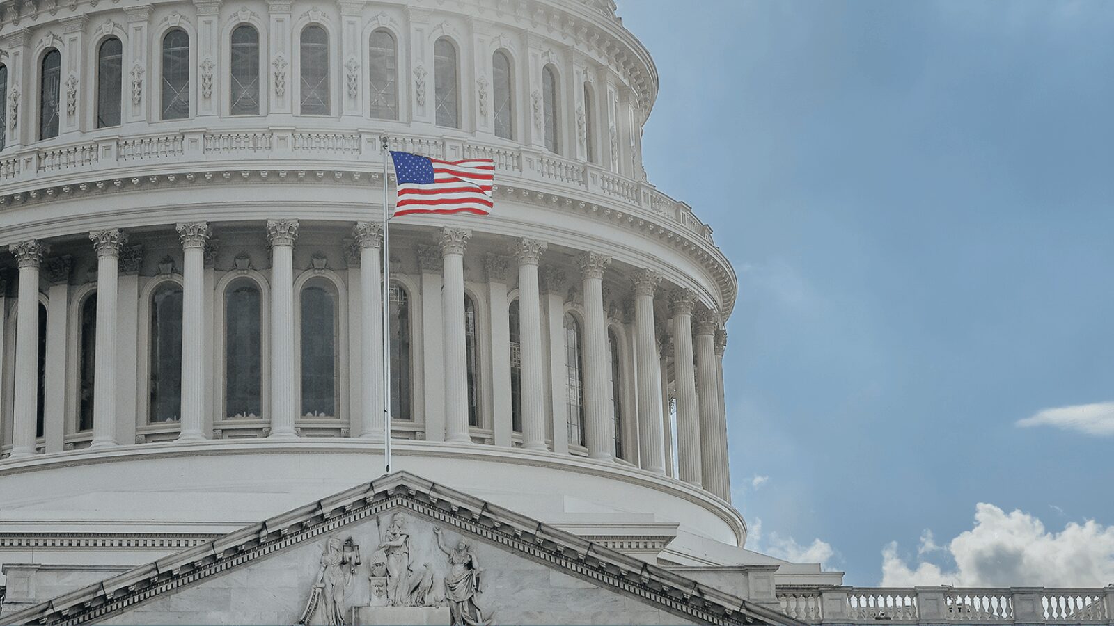 The American flag waves in front of the U.S. Capitol building, featuring its distinctive dome and classical columns against a partly cloudy sky