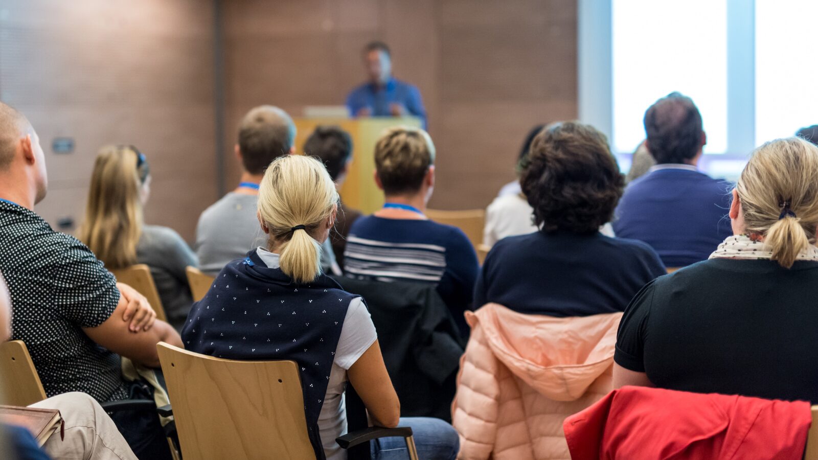 Audience members seated in a conference room, attentively listening to a speaker presenting at a podium in the front of the room