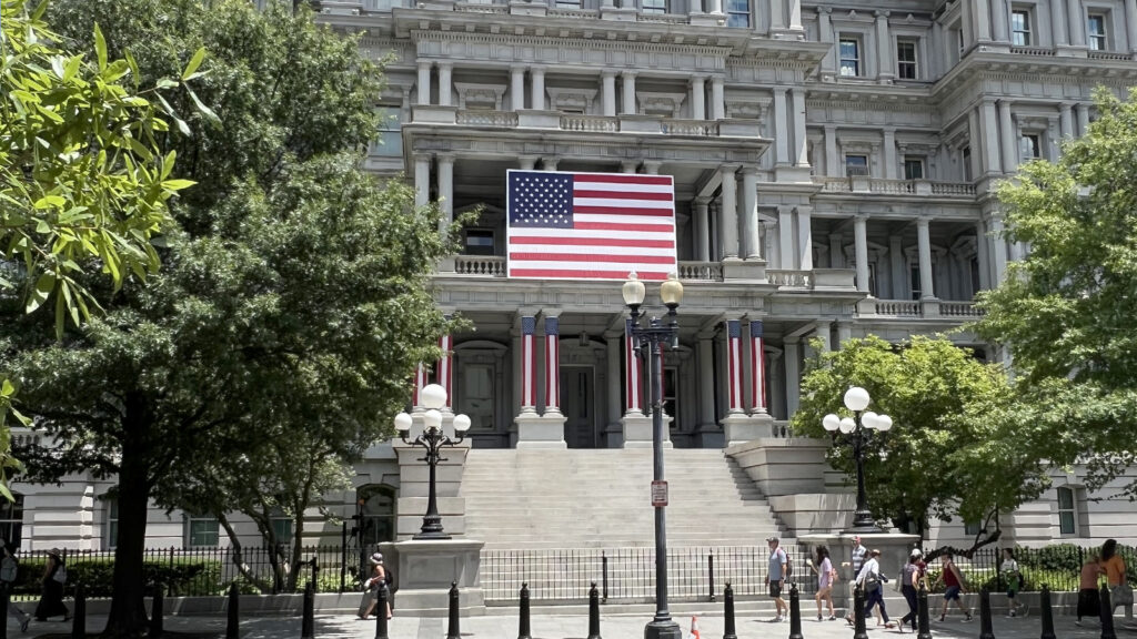 Historic government building with an American flag and patriotic banners, surrounded by trees and street lamps, with people walking by_1600x900