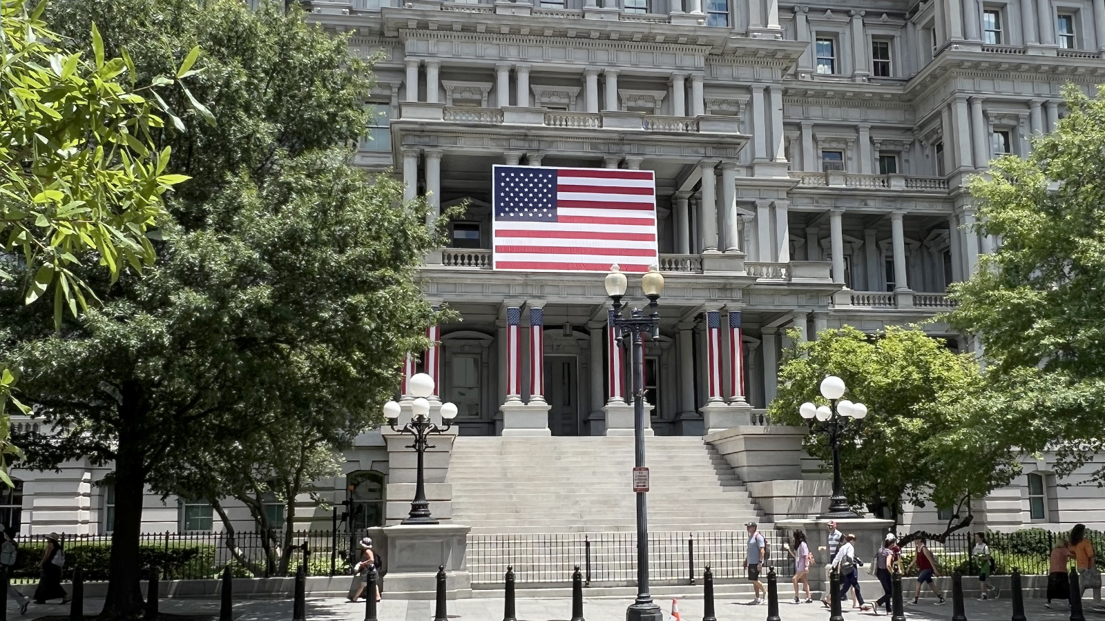 Historic government building with an American flag and patriotic banners, surrounded by trees and street lamps, with people walking by_1600x900