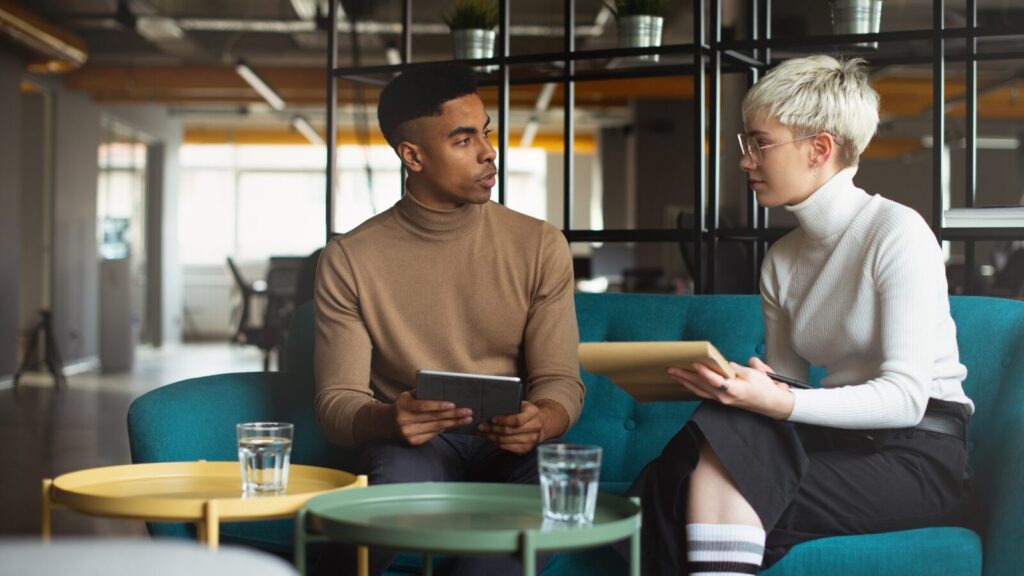 Two young professionals engaged in a discussion in a modern office lounge, holding a tablet and folder, with water glasses on tables