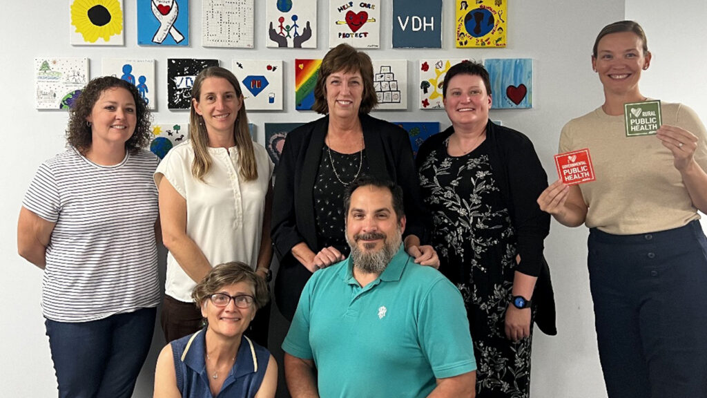 A group of seven public health professionals posing together, with colorful public health-themed artwork displayed on the wall behind them