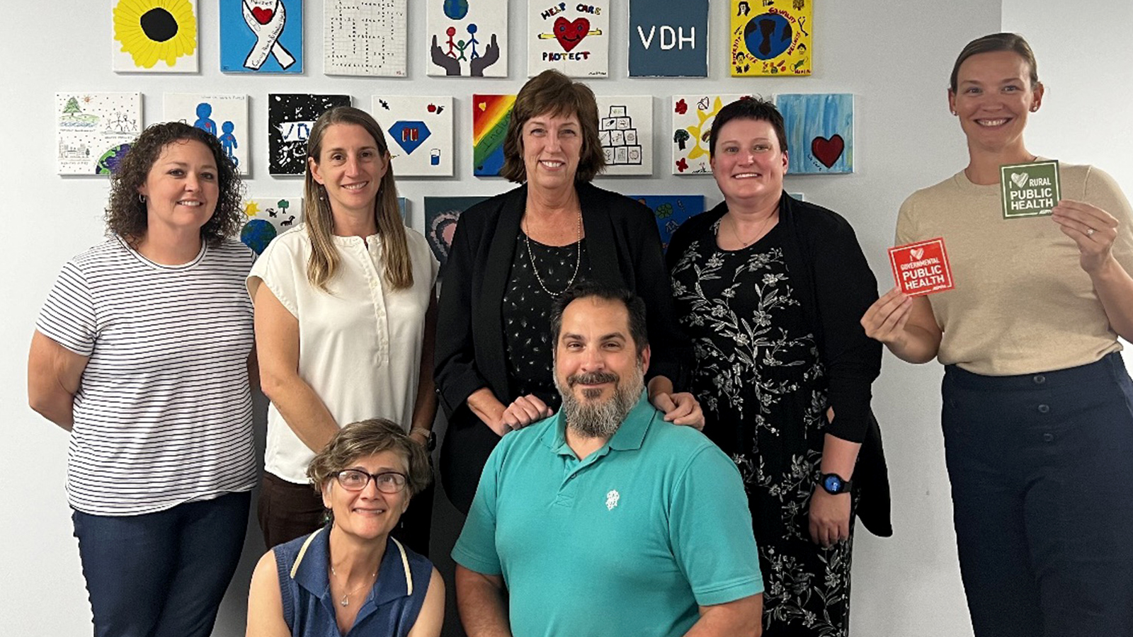 A group of seven public health professionals posing together, with colorful public health-themed artwork displayed on the wall behind them