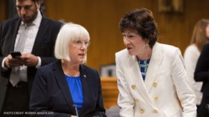Senators Patty Murray and Susan Collins having a conversation during a Senate session, with another person in the background checking their phone.