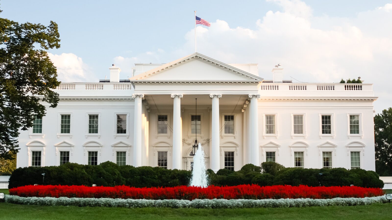 The White House North Portico with an American flag flying on top, framed by blue skies, lush greenery, and a red flower bed in the foreground