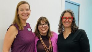 Three women smiling together during the ASPPH visit to the University of Kentucky College of Public Health. Two are wearing glasses, and all are standing indoors.