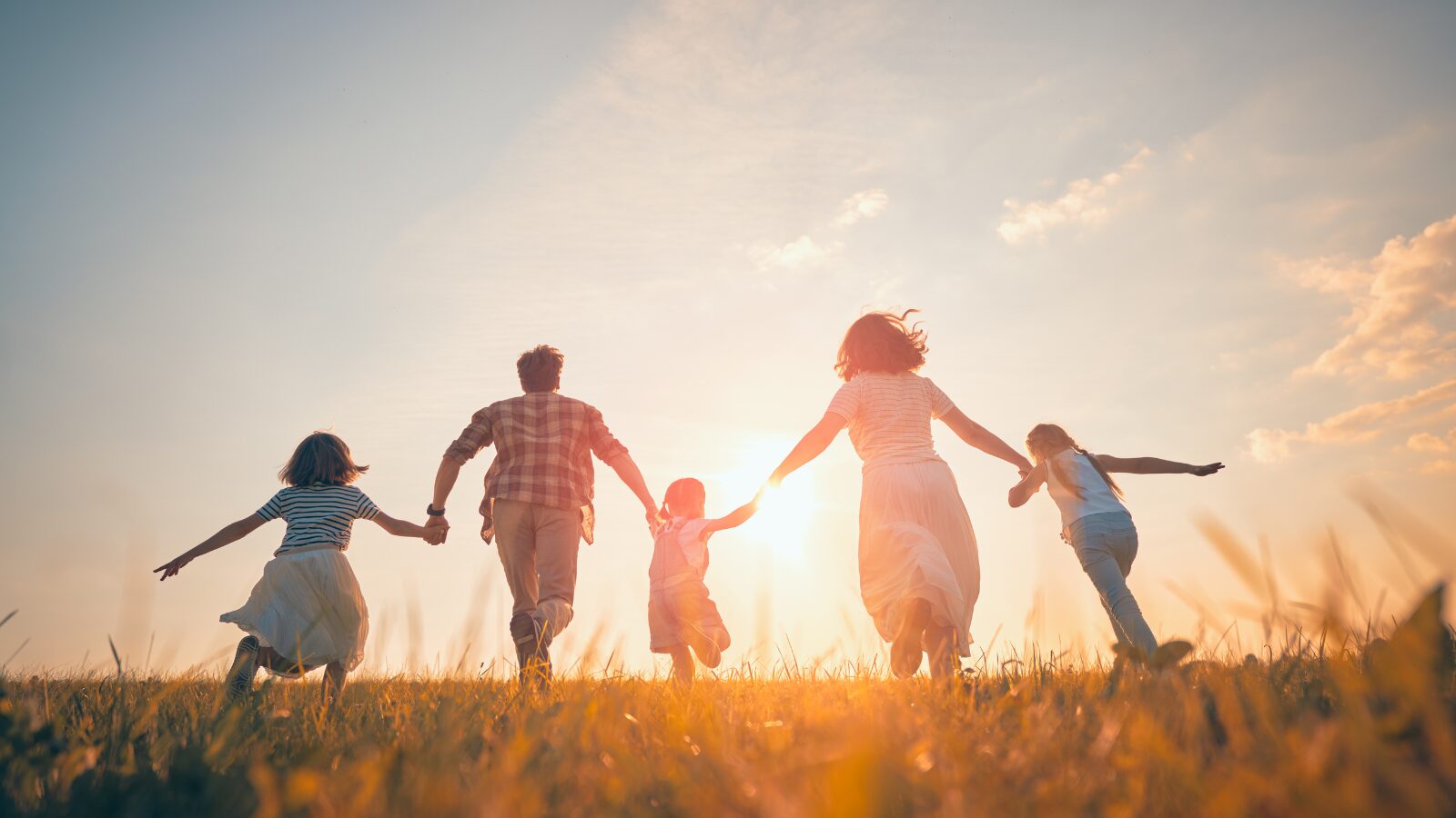 A family of five, including two adults and three children, running through a sunlit grassy field, holding hands and smiling against a warm, bright sky