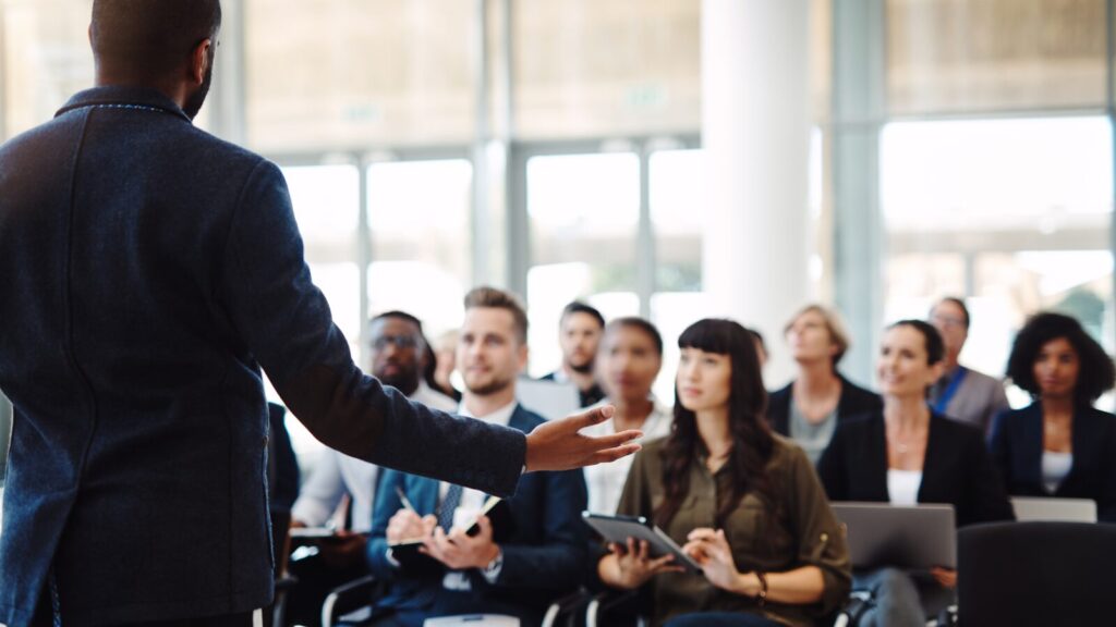 Speaker addressing a diverse audience in a modern conference setting with attendees listening taking notes and using laptops or tablets