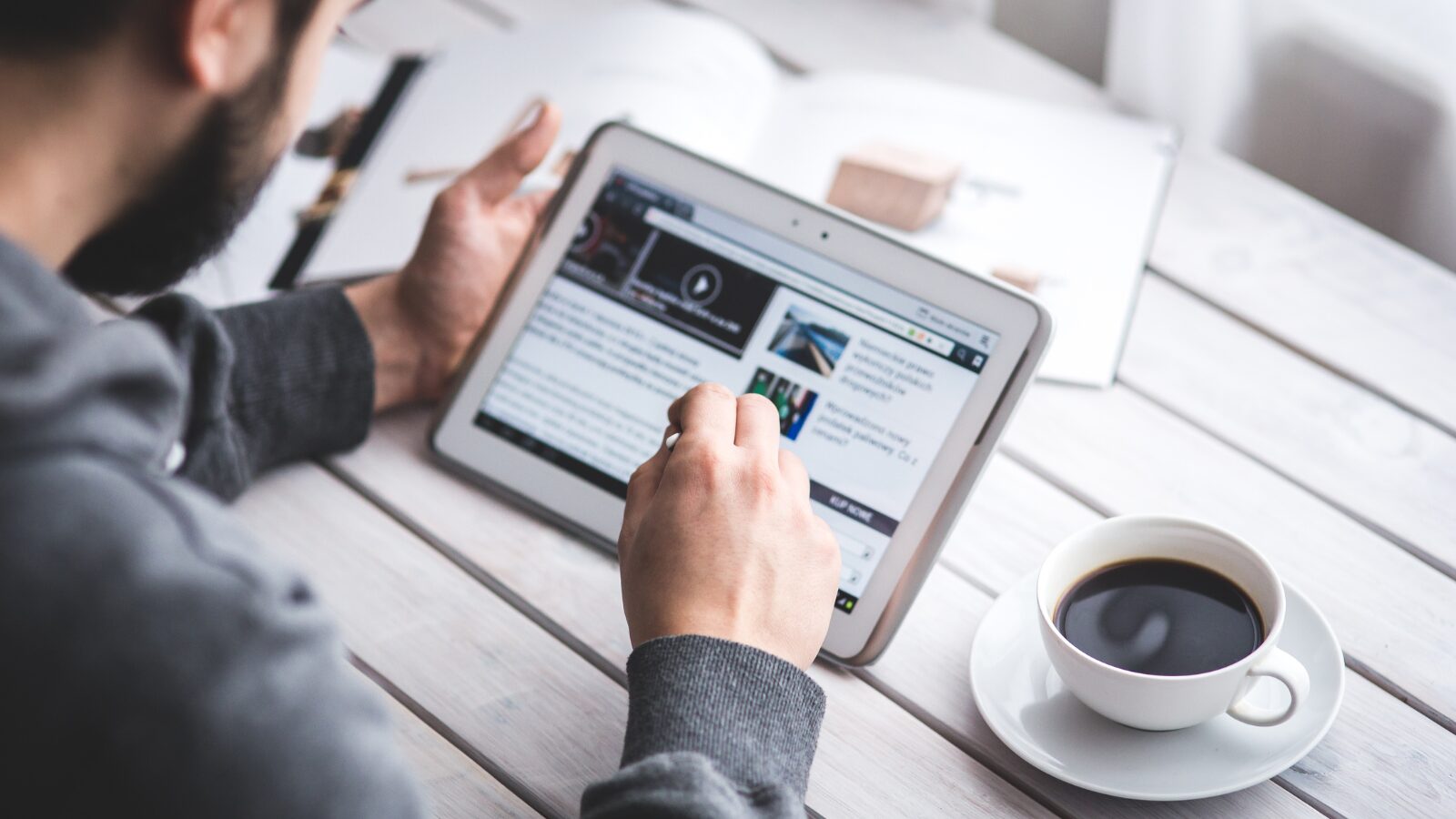 Person using a tablet with a stylus to browse online content while sitting at a white wooden table with a cup of black coffee