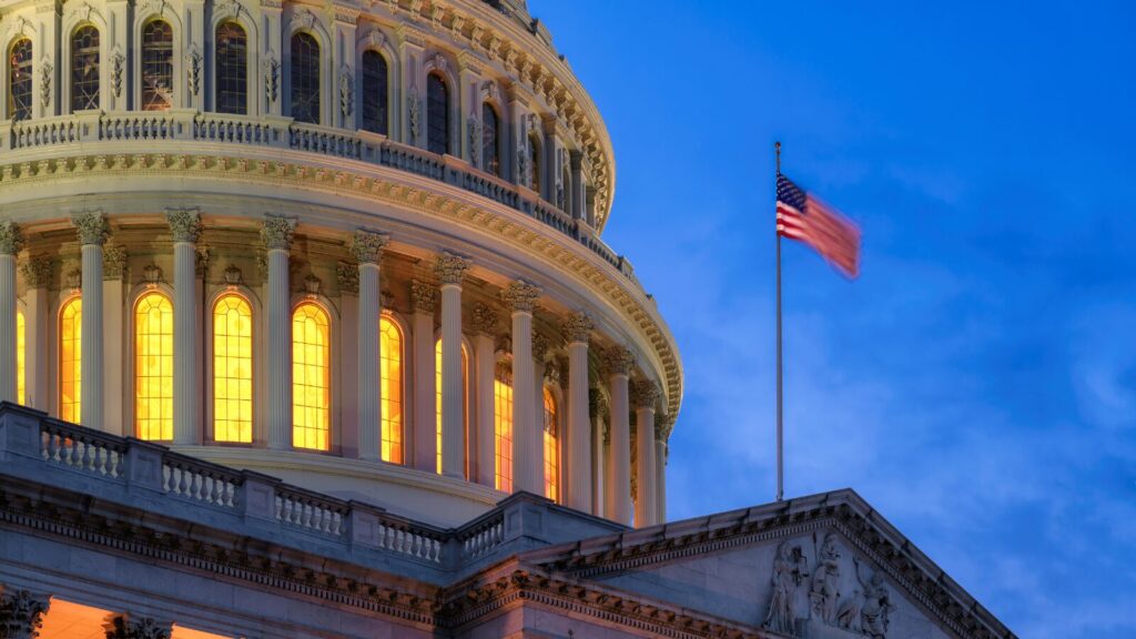 Close-up of the US Capitol dome at dusk with glowing windows white columns and an American flag waving against a deep blue sky