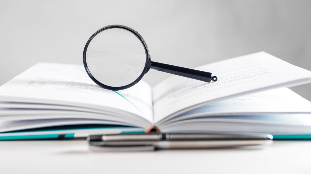 Magnifying glass resting on an open book with two pens in the foreground, symbolizing research, analysis, or investigation