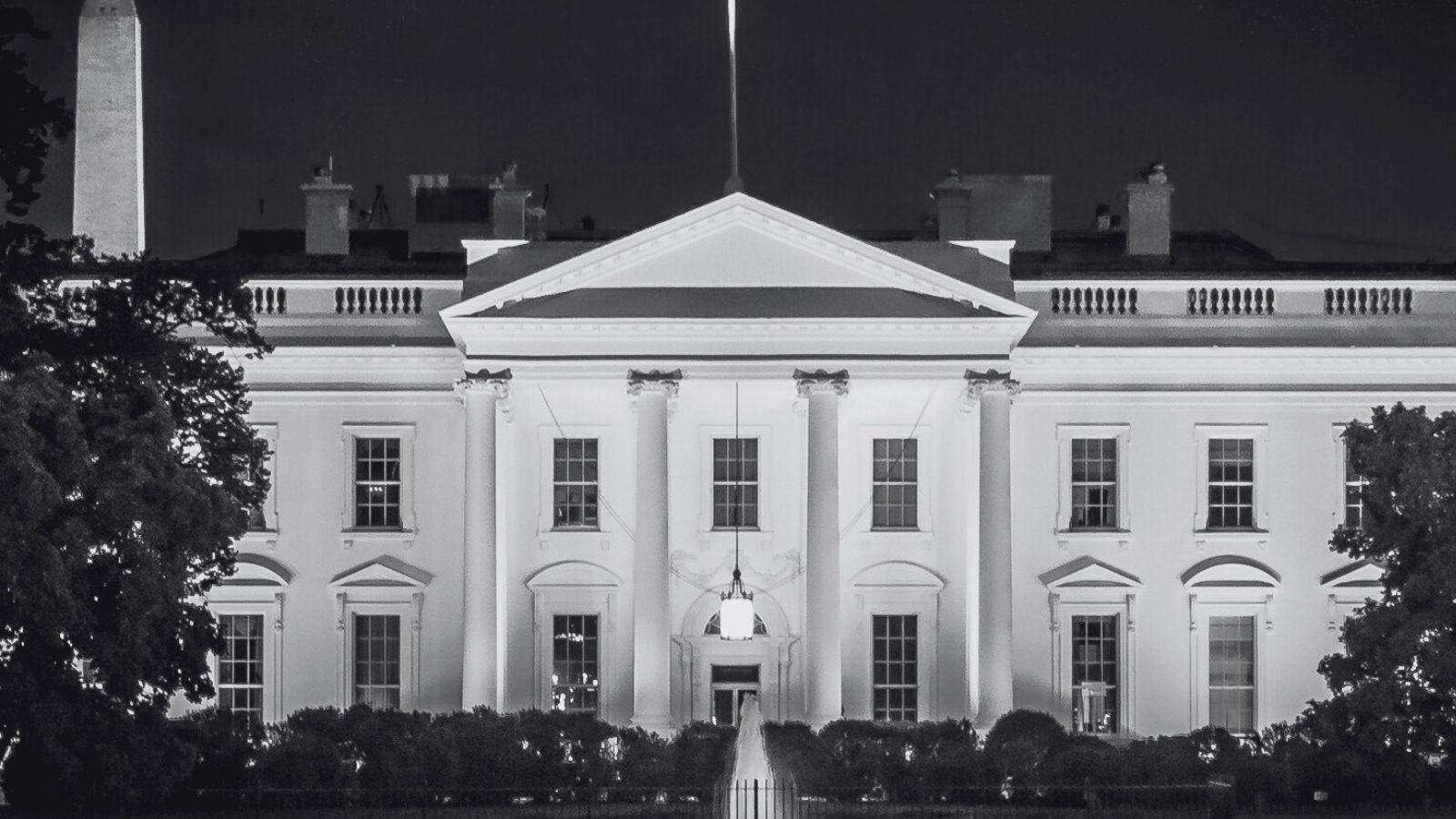 Black and white photo of the White House at night with lights illuminating the building and trees partially framing the scene