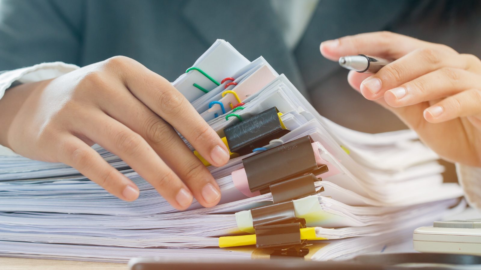 Close-up of hands sorting through stacks of documents clipped together with colorful paper clips and binder clips while holding a pen
