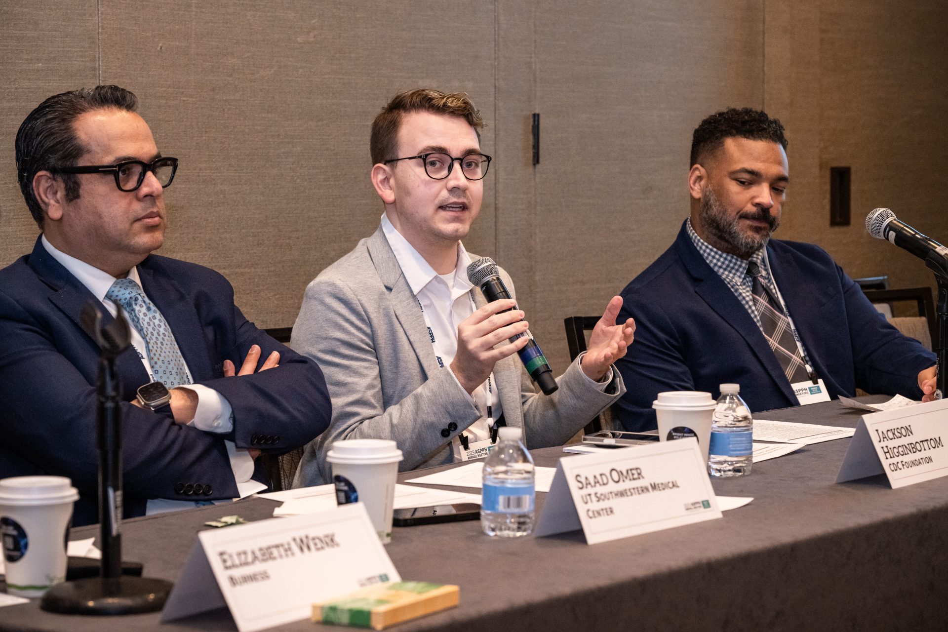 Three public health professional seated at a table while one speaks into a microphone