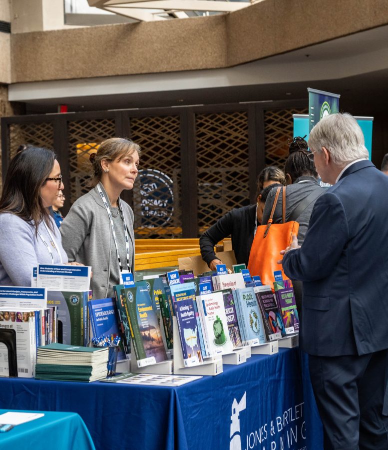 A man approaches a table full of books and speaks with two women standing behind the table