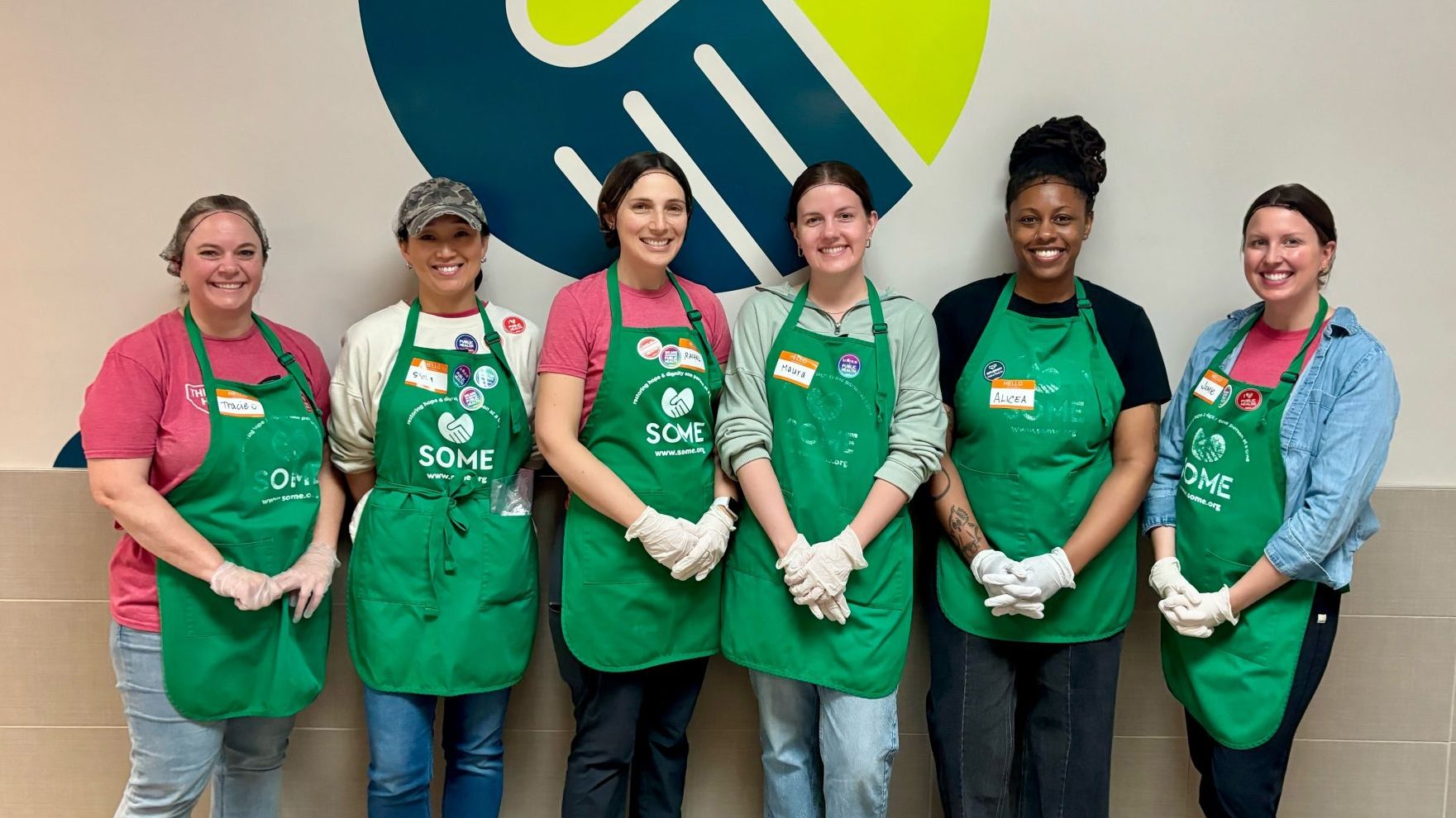 Six people in food kitchen wearing green aprons with "SOME" written on them