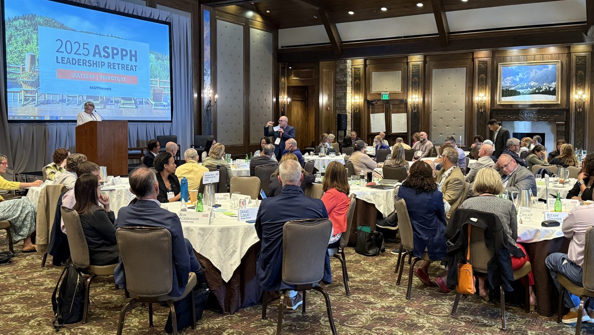Groups of people sitting at tables as someone speaks at a podium